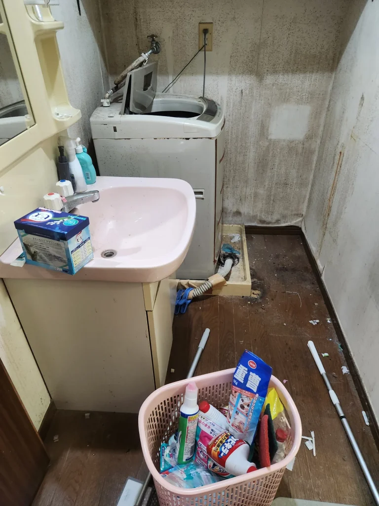 洗面台と洗濯機まわりに置かれた掃除用品と汚れた壁面